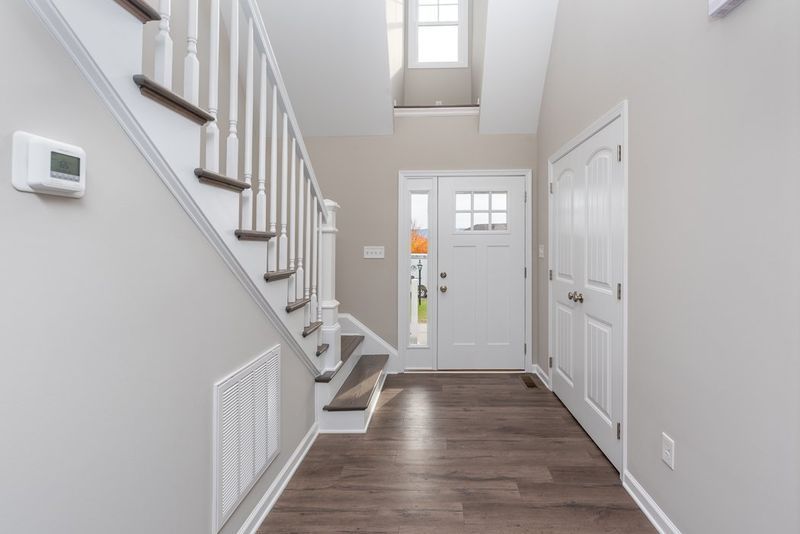 Entryway with staircase, front door, and white trim; light gray walls and dark wood-look flooring.