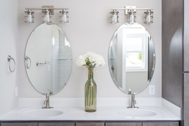 Two oval mirrors above bathroom sinks with silver fixtures, flower vase in between.