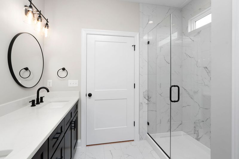 Modern bathroom with black accents: vanity, mirror, shower, and door. White walls and marble-like tile.