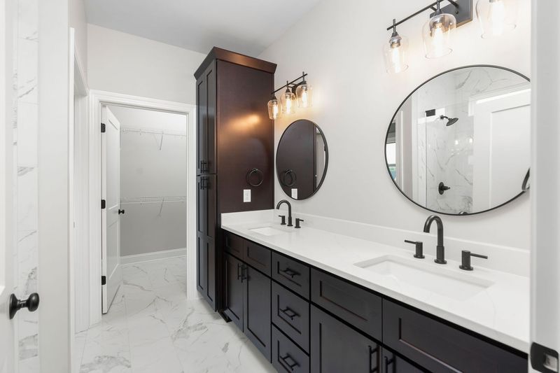 Bathroom with dark cabinets, white countertops, two sinks, round mirrors, and a tall storage cabinet.