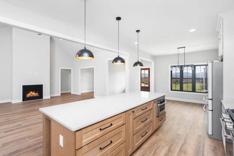 Spacious kitchen with island and light wood cabinets, leading to a living area with a fireplace and view.