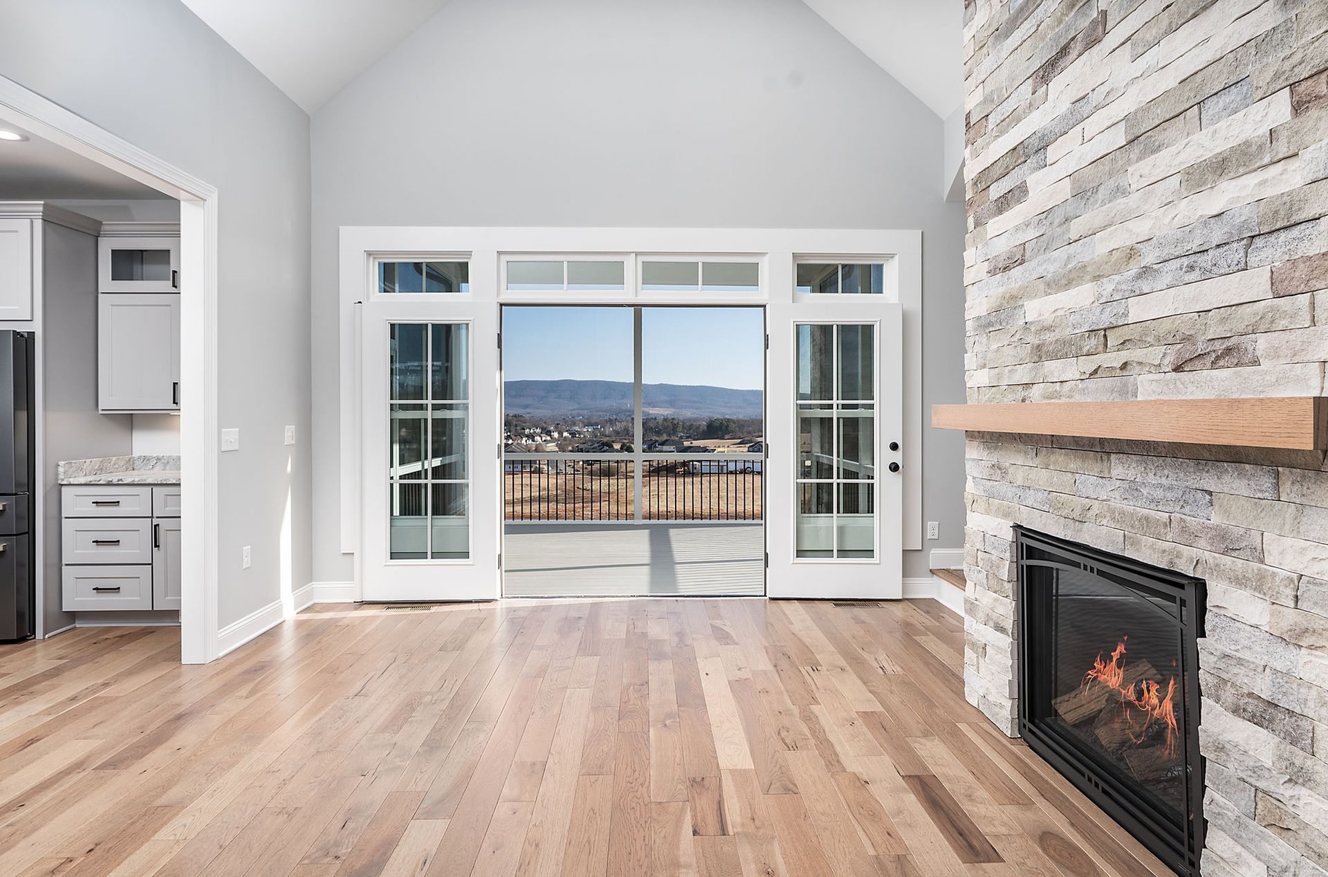 Empty living room with wood floors, stone fireplace, and large sliding glass doors overlooking a landscape.