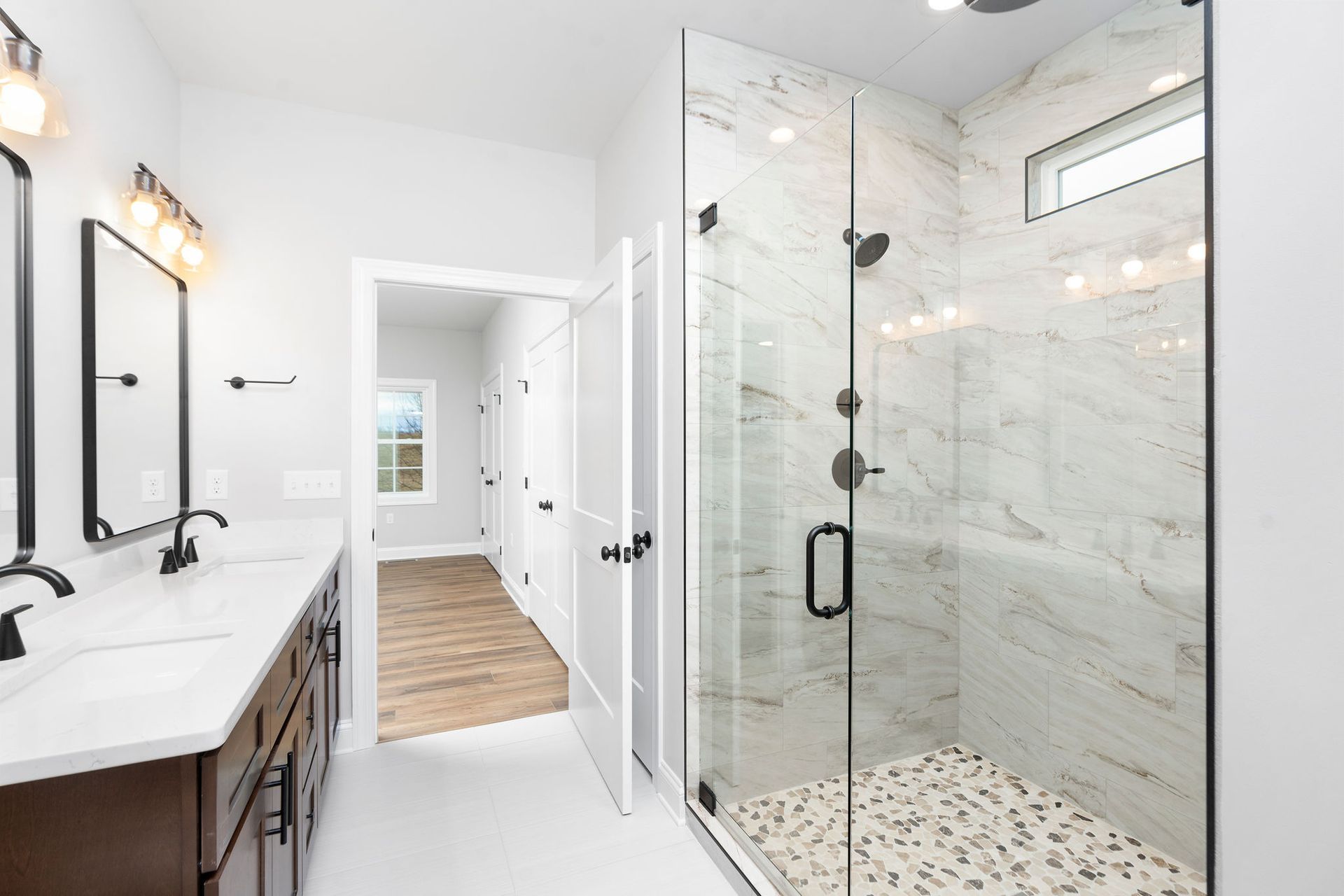 Modern bathroom with a glass shower, double vanity, and open doorway. White walls, gray floor.