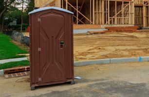 A brown portable toilet is sitting in front of a house under construction.