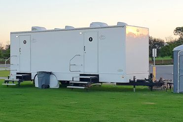 A white trailer is parked in a grassy field next to a blue portable toilet.