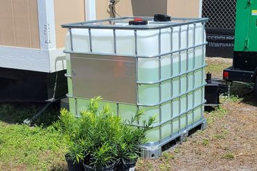 A large plastic container filled with liquid is sitting on a pallet next to potted plants.