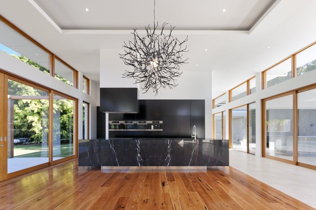 Modern kitchen with a black marble island and chandelier, surrounded by floor-to-ceiling windows and wood floors.