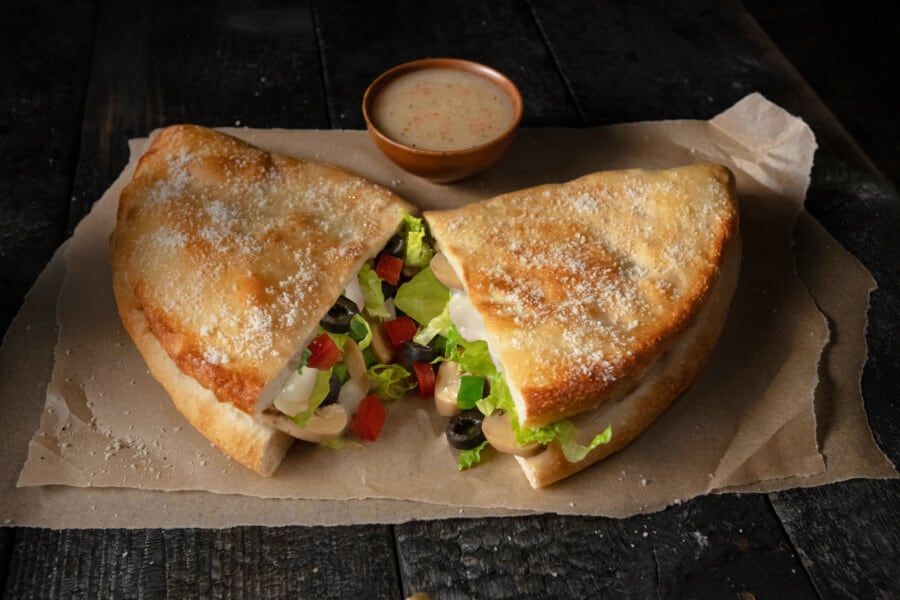 A sandwich with lettuce , olives , tomatoes and cheese is cut in half on a wooden table.