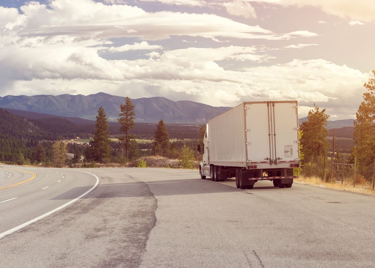 A semi-truck parked on the shoulder of a highway with scenic mountains and trees in the background under a cloudy sky.