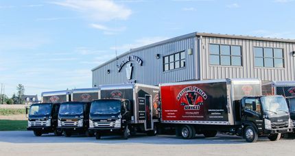 Four delivery trucks were parked in front of a large metal warehouse on a sunny day.