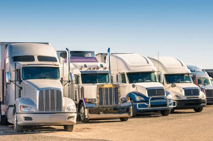 A row of several semi-trucks parked in a gravel lot under a clear blue sky.