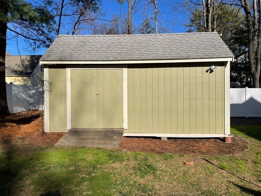 Small beige garden shed with a gray shingled roof on a grassy backyard.