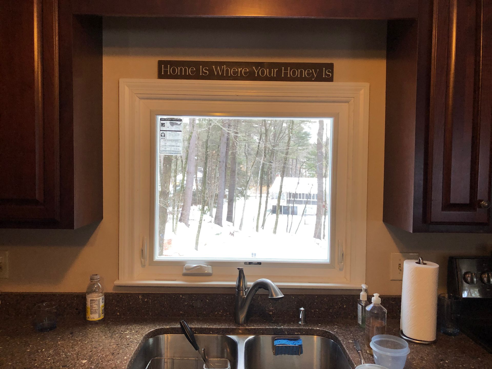 Kitchen window above sink overlooking snowy forest, with dark wood cabinets and granite countertop.