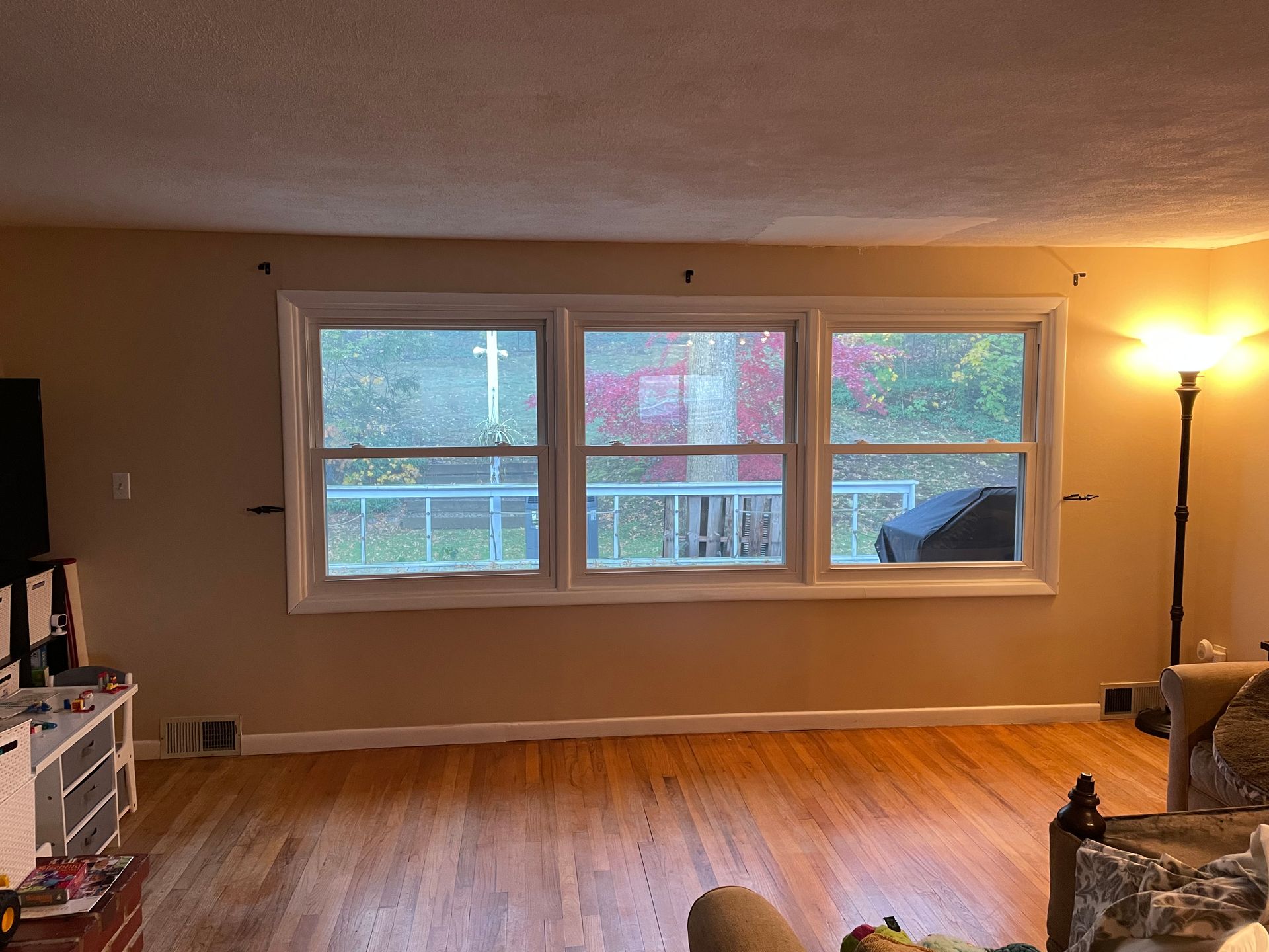 Empty living room with hardwood floors, large windows, and a lit floor lamp