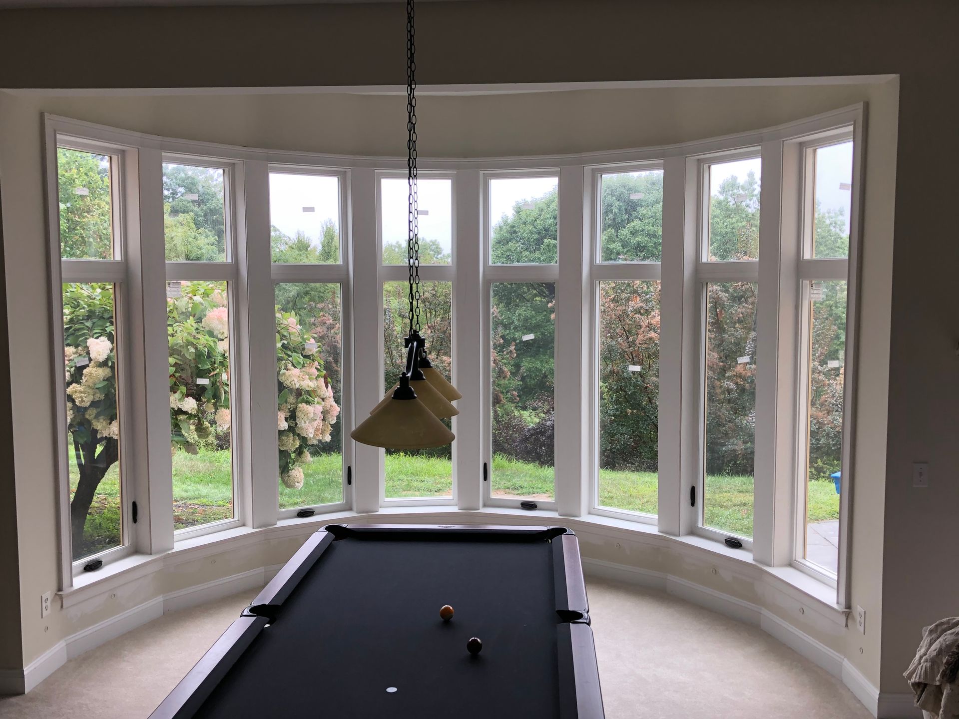 Curved bay windows overlooking greenery, with a black pool table and hanging light in a bright room