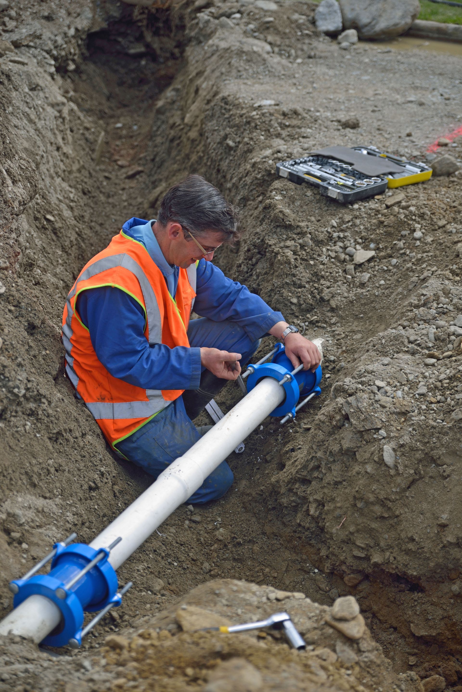Worker in orange vest kneels in trench, connecting white pipe with blue fittings.