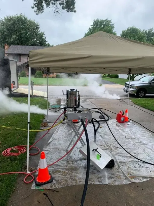 A machine under a tent emits steam onto a driveway.  Hoses, a cone, and red containers are nearby.