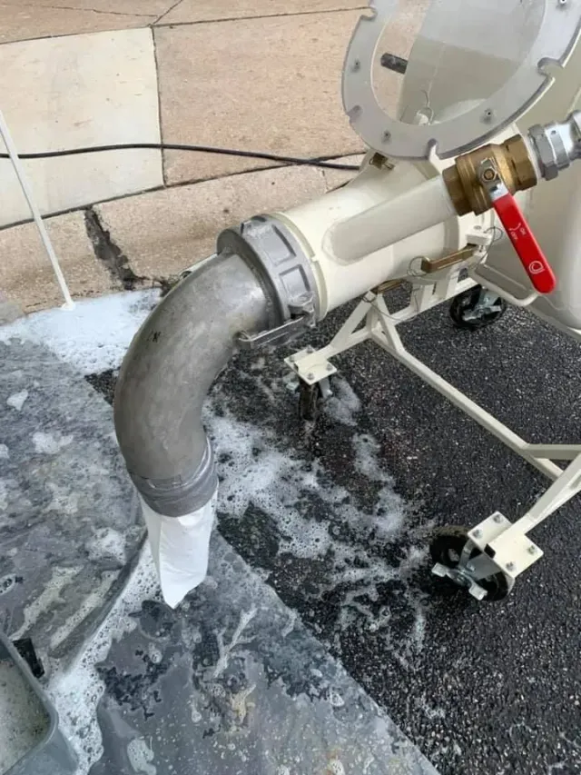 A white machine with a metal pipe is expelling foamy liquid onto a dark surface.