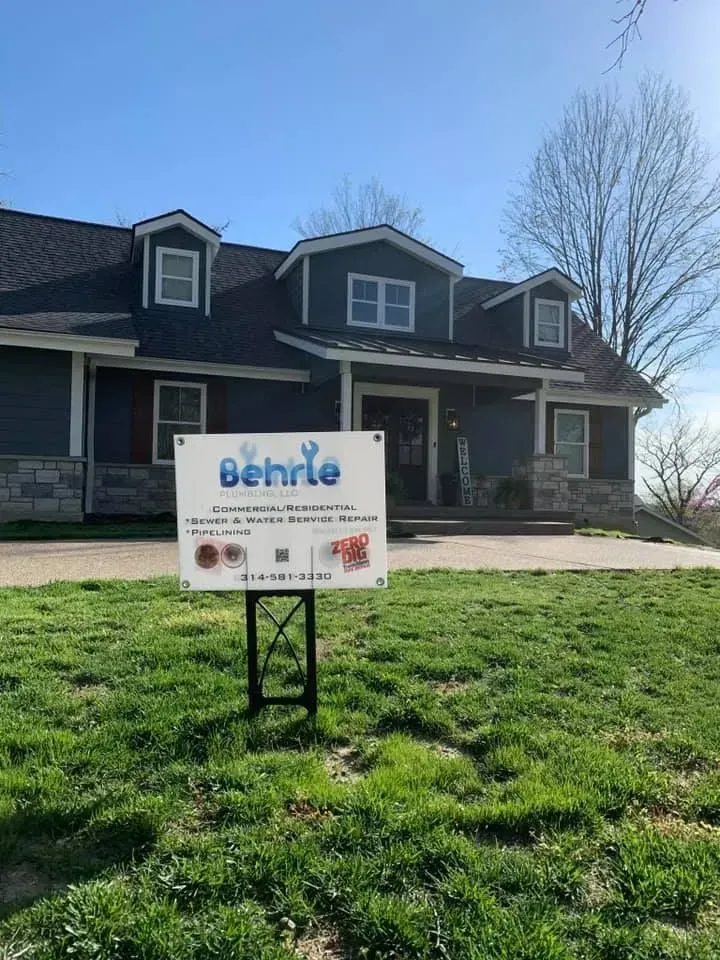 Sign for Behrie, in front of a blue house with dormers and stone accents, on a grassy lawn under a clear sky.
