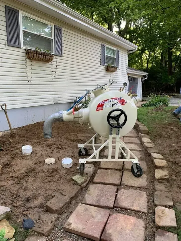 A white machine on wheels next to a house with a dirt area, pathway of stones, and a shovel.