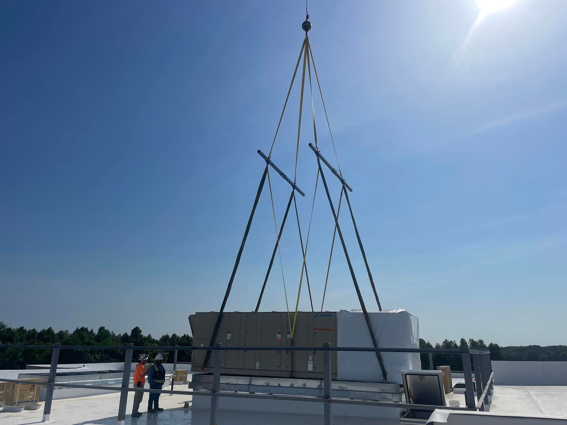 A crane lifting an HVAC unit onto a flat roof, two workers observing under a bright blue sky.