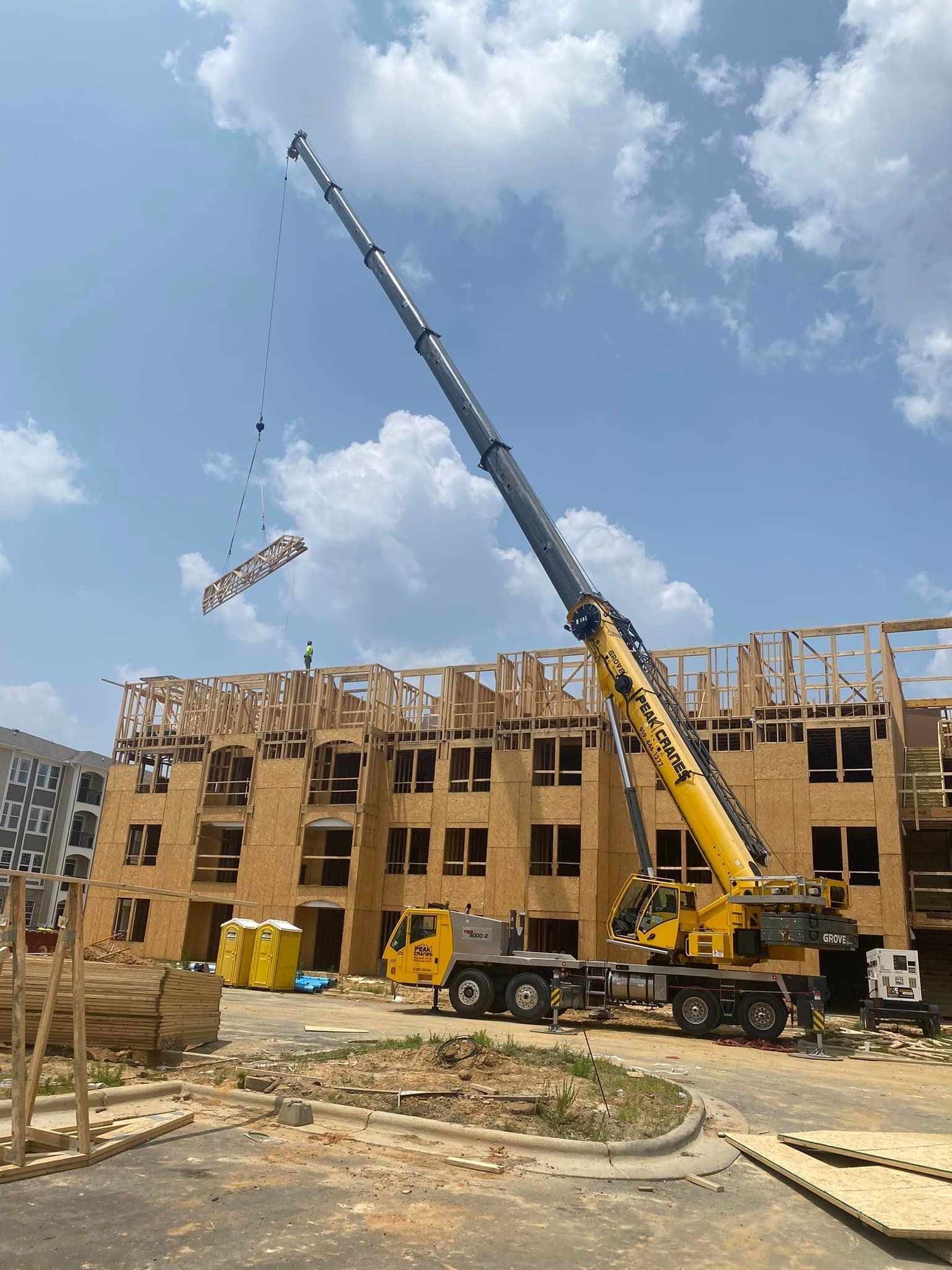 A large yellow crane lifts wood beams onto a multi-story building under construction on a sunny day.