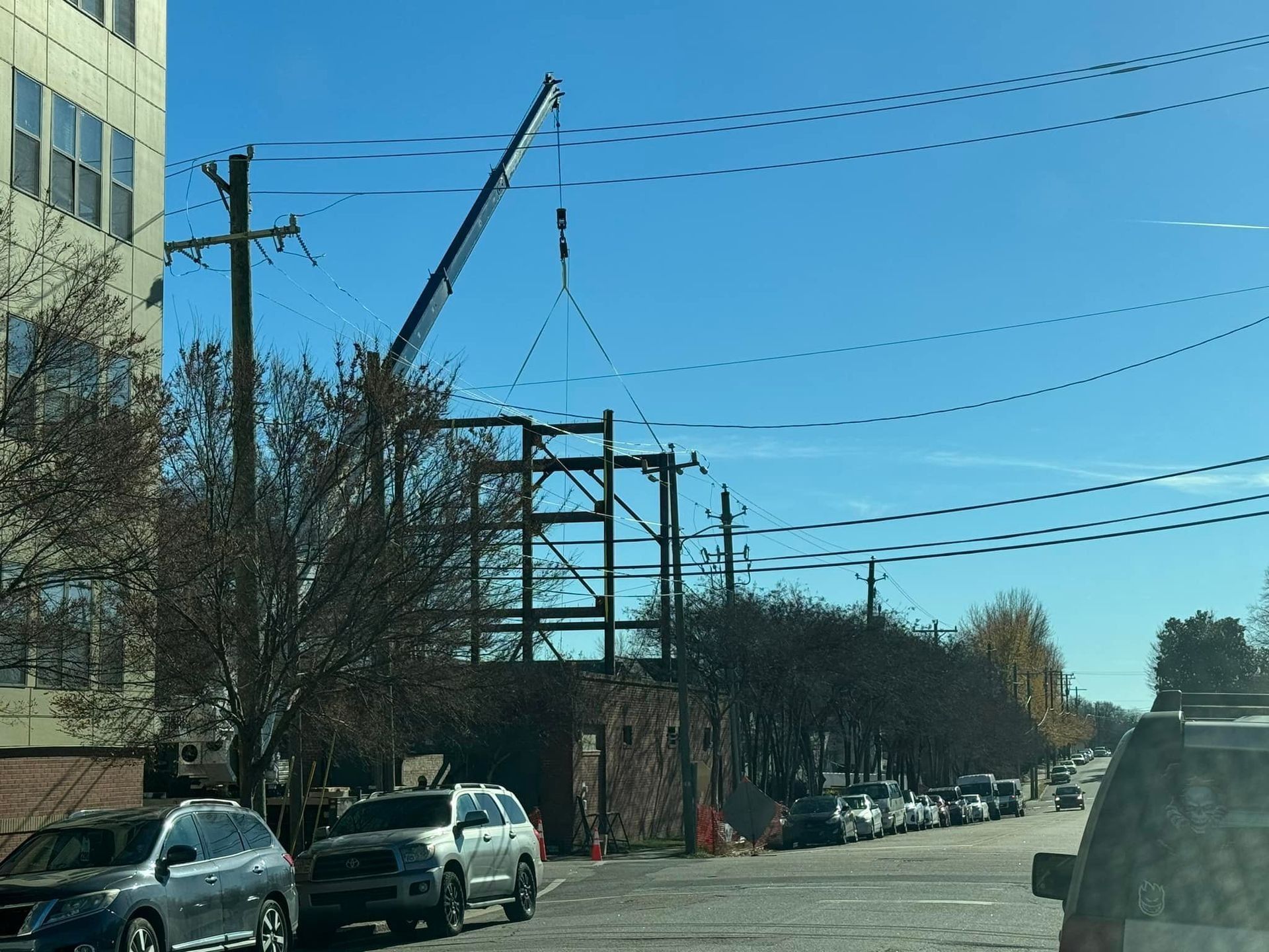 Construction site with a crane lifting steel beams; cars parked on a street; a blue sky.