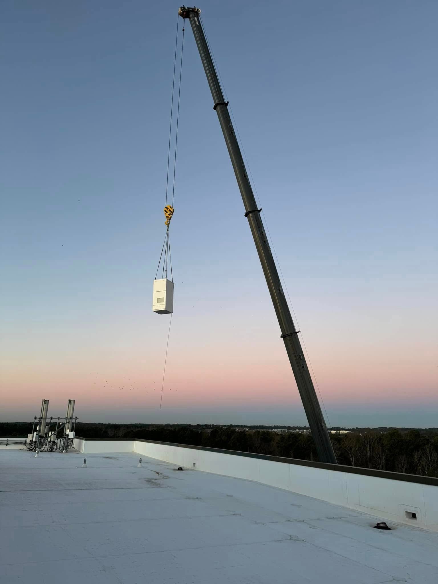 Crane lifting a white box onto a rooftop at sunset. Blue, pink, and orange sky.
