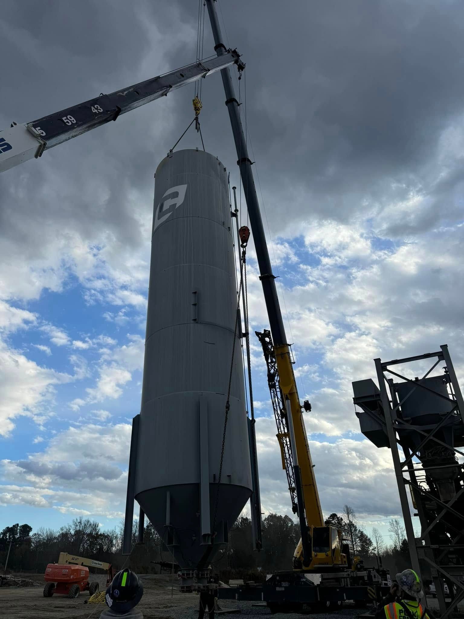 Two cranes lifting a tall, gray industrial silo at a construction site under a cloudy sky.