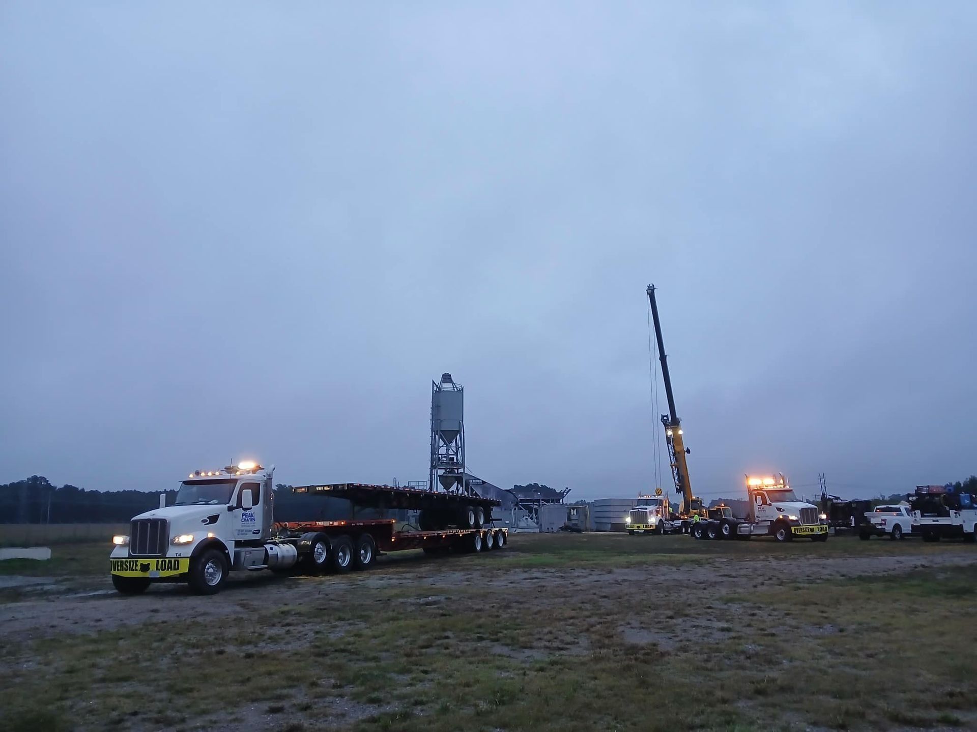 A flatbed truck, crane, and other vehicles at a construction site under a cloudy sky.