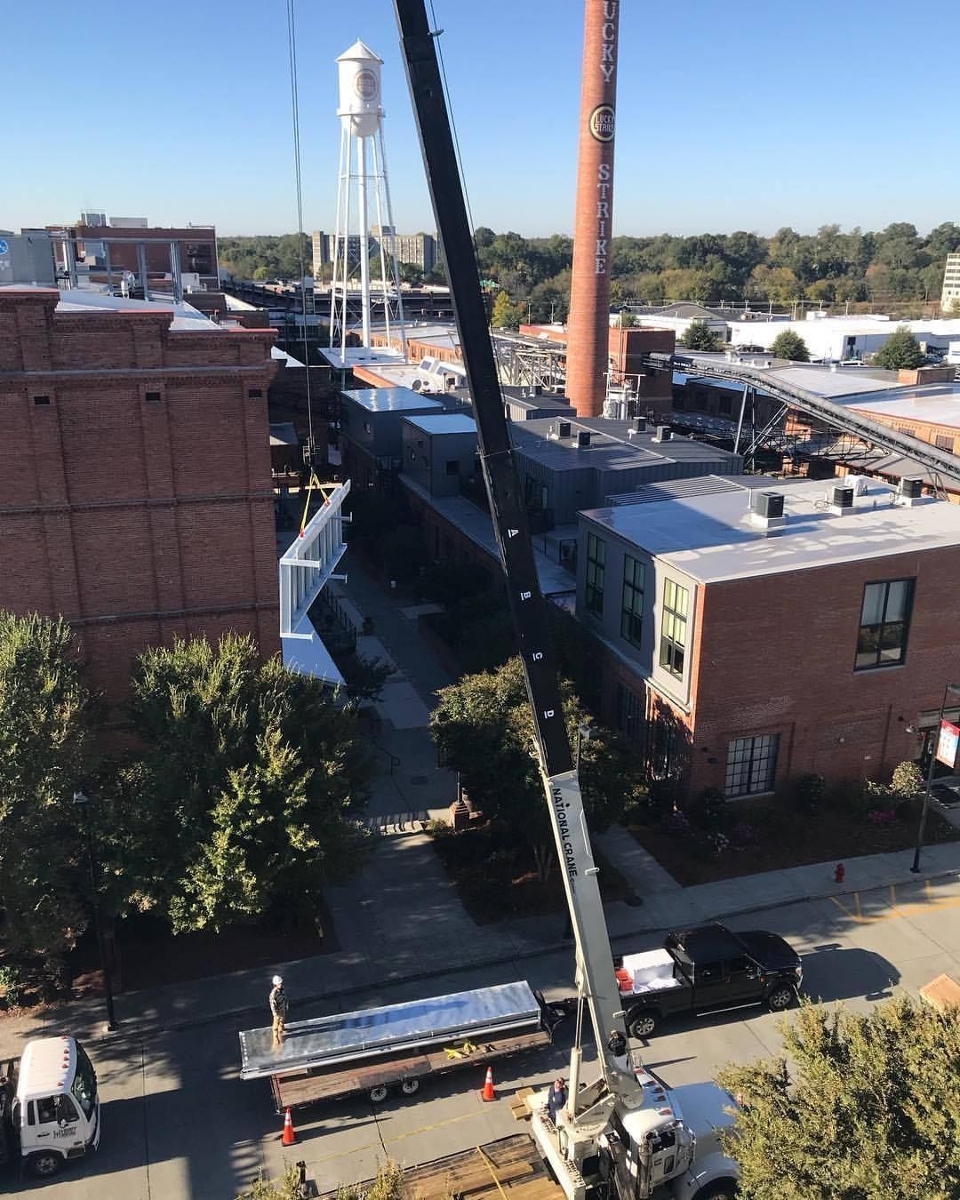 Crane lifting a long, silver structure onto a building under construction, between brick buildings, near a water tower.