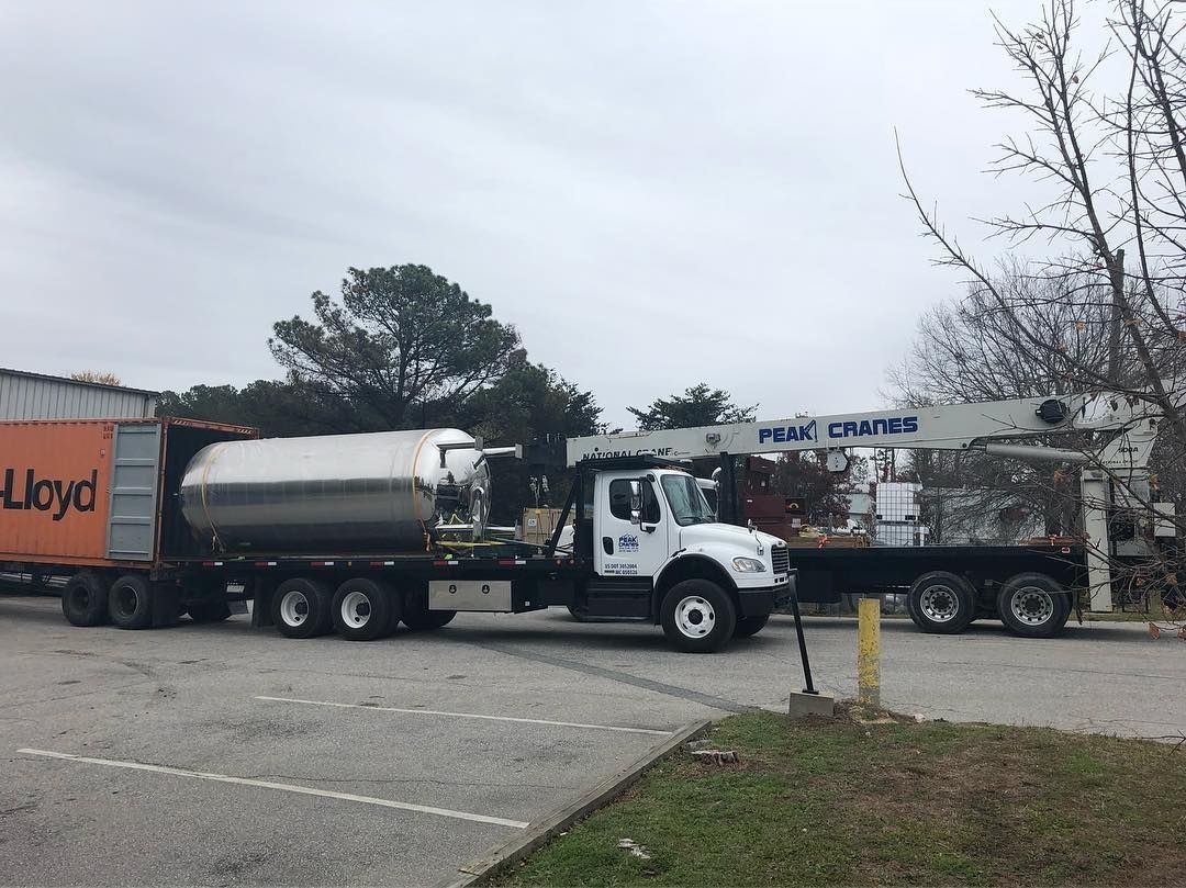 White tanker truck with container trailer, parked beside a crane truck, gray overcast sky.