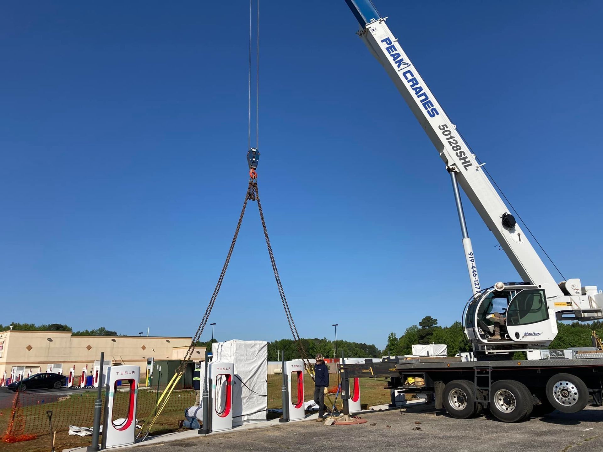 Crane lifting a charging station component at a Tesla Supercharger site. Clear sky, construction in progress.