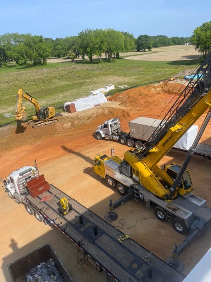 Construction site with a yellow crane and excavator; trucks and dirt. Sunny day with trees in the background.