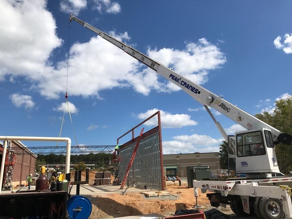 Crane lifting a metal wall panel at a construction site on a sunny day with blue sky and some clouds.