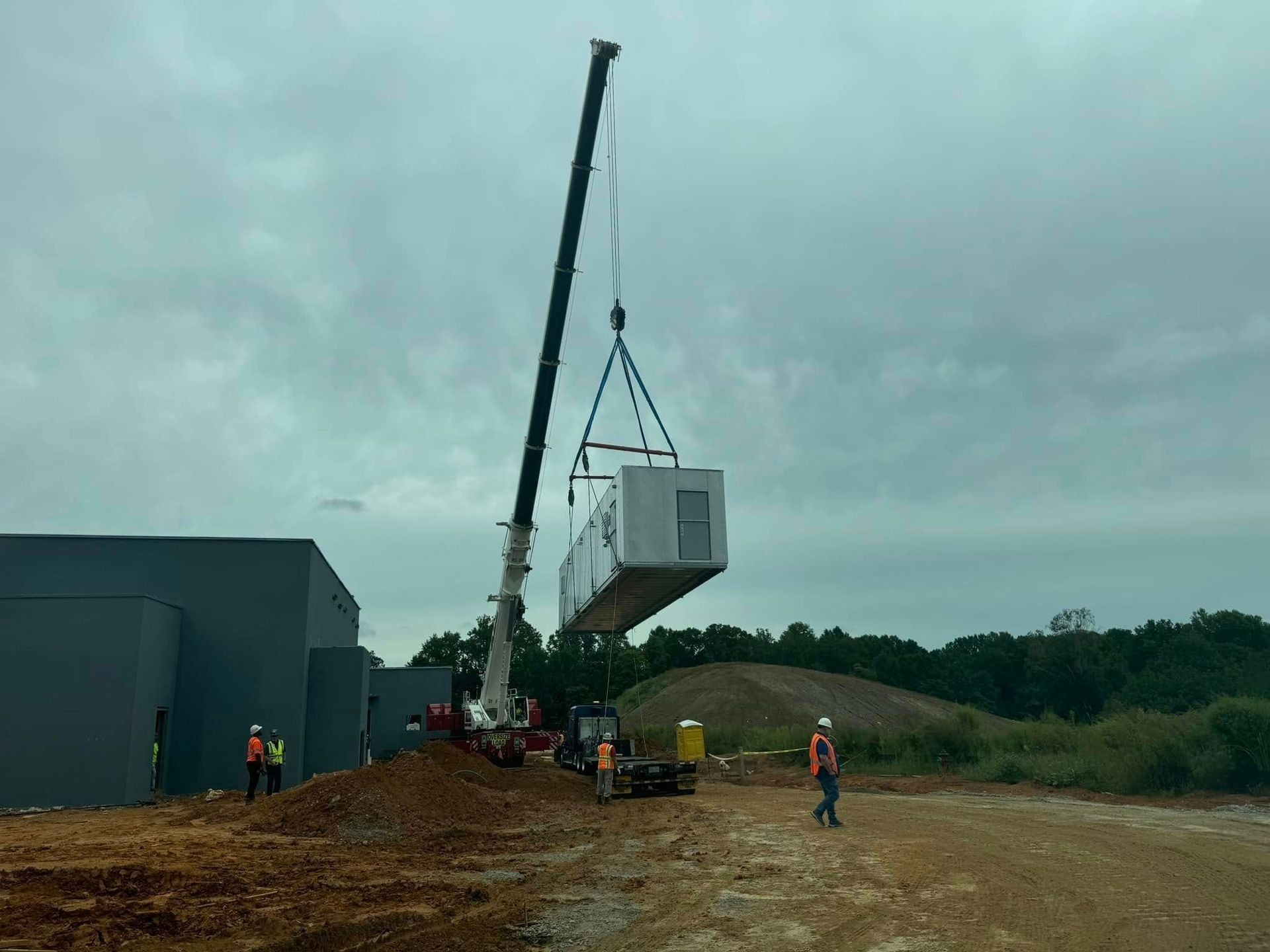 A crane lifting a modular building component near a gray structure and construction site. Workers in safety vests are present.