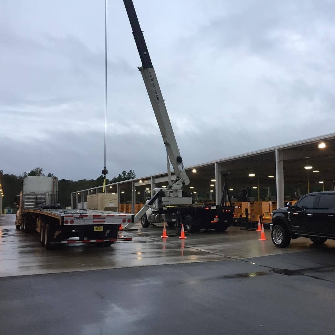 Crane lifting a large object from a flatbed truck near a building with a covered walkway; gray overcast sky.