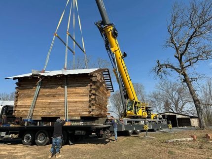 Crane lifting a wooden cabin off a flatbed trailer in a grassy field on a sunny day.