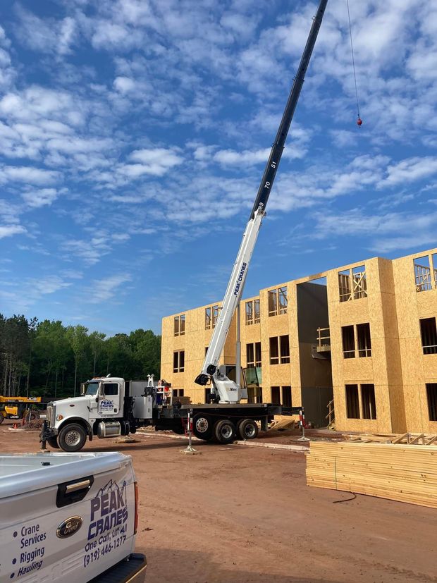A large crane lifting materials at a construction site, near a partially built wooden building, under a blue sky.
