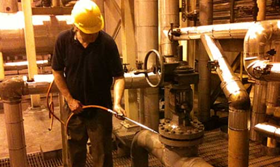 A worker in a yellow hard hat using a hose in an industrial plant room with pipes and valves