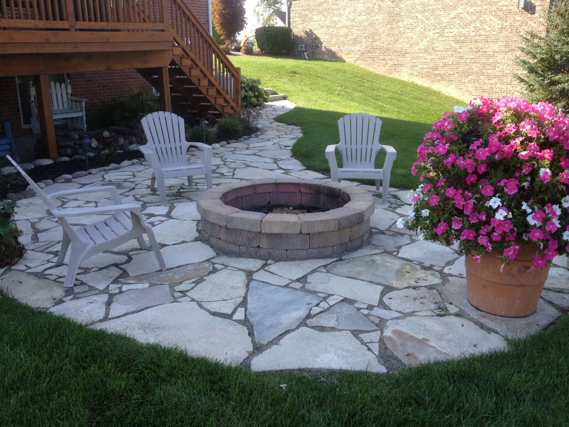 A fire pit is surrounded by chairs and flowers on a stone patio.