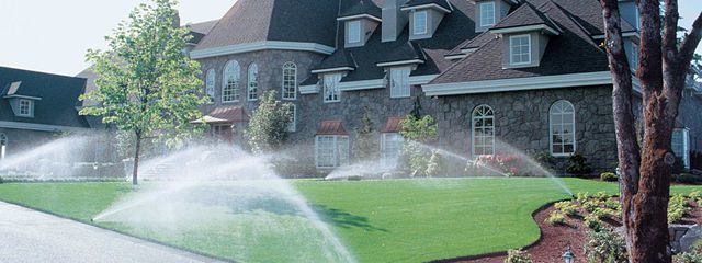 A lawn sprinkler is spraying water on a lush green lawn in front of a large house.
