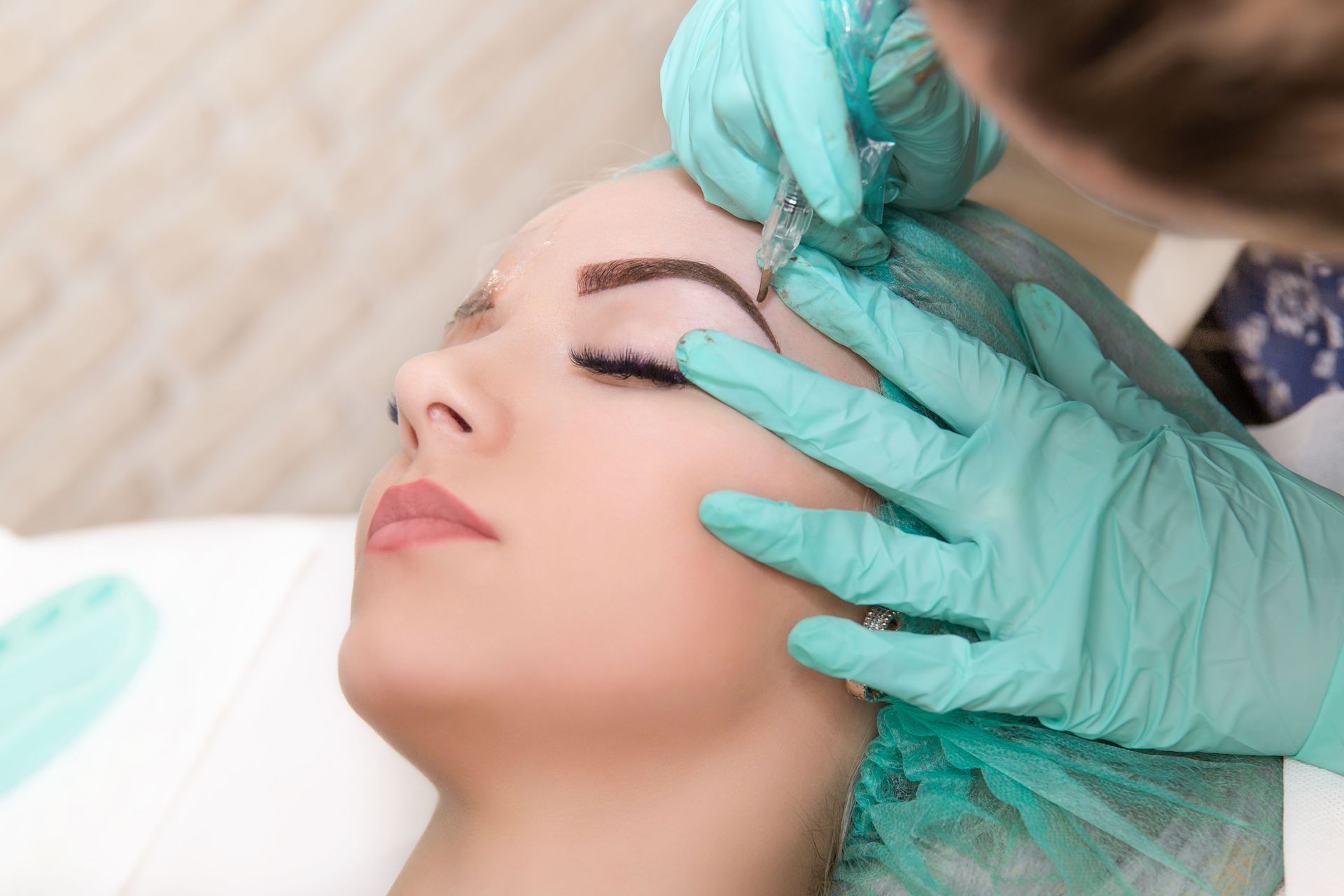 Woman receiving eyebrow microblading treatment. A technician in teal gloves and cap holds a tool, creating a new brow shape.