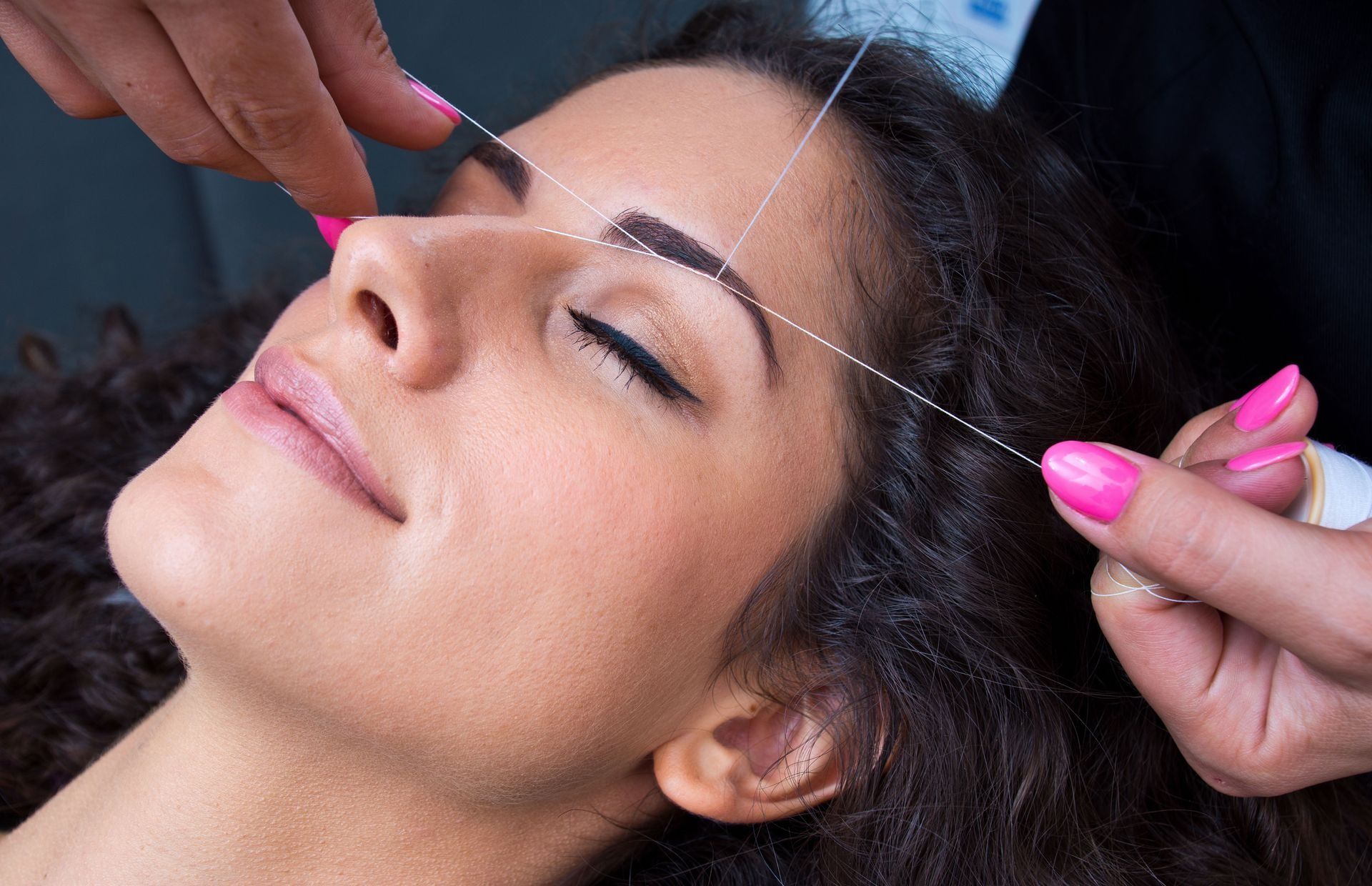 Woman having her eyebrows threaded. Hands manipulating thread near her face; she has closed eyes.