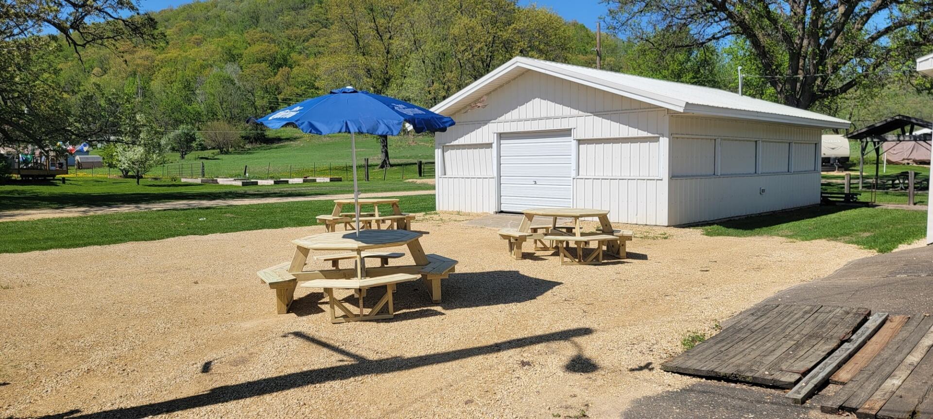 A white shed with a blue umbrella and picnic tables in front of it.