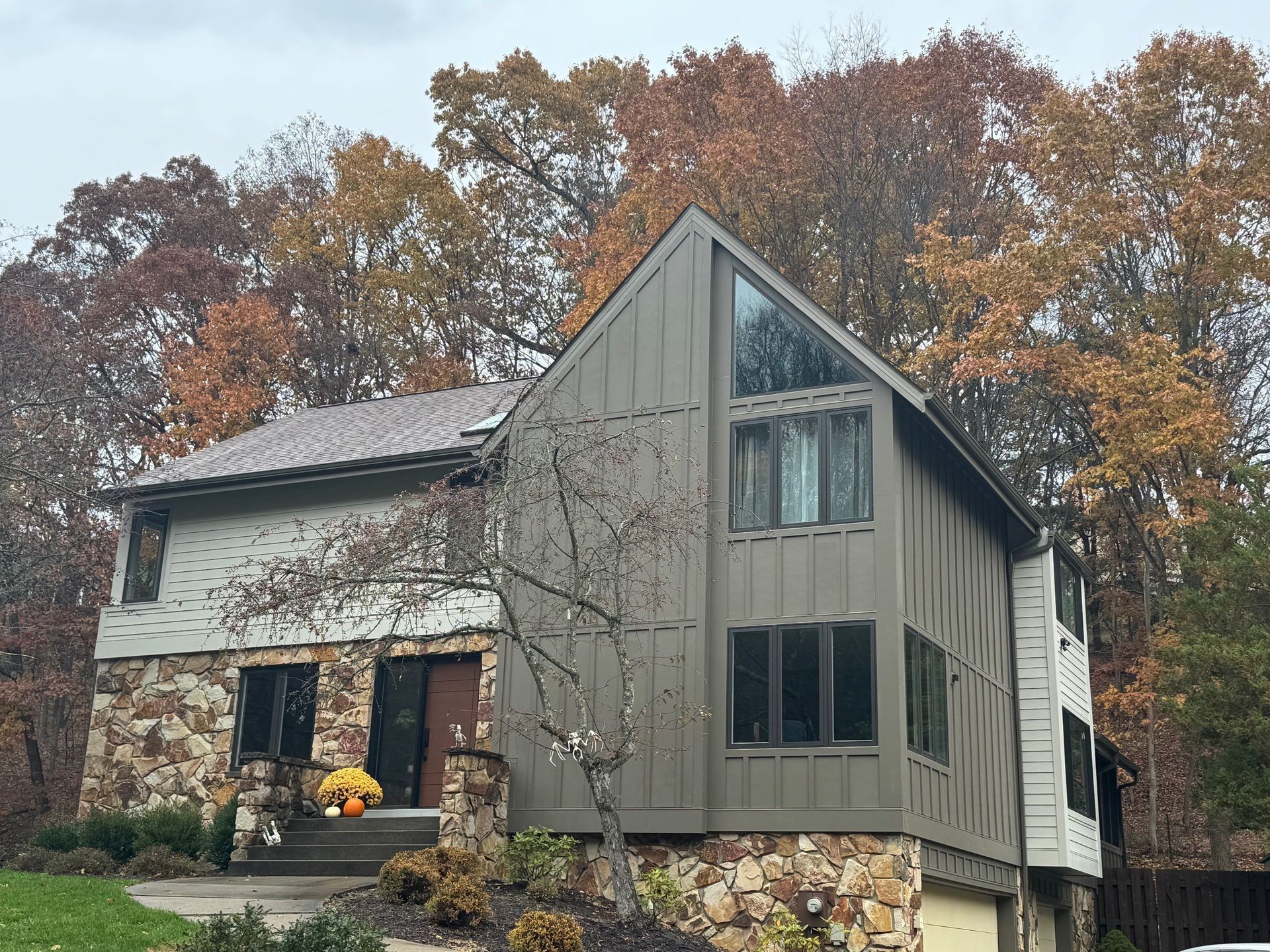 Two-story house with stone facade, dark brown siding, and a large triangular window, surrounded by fall trees.