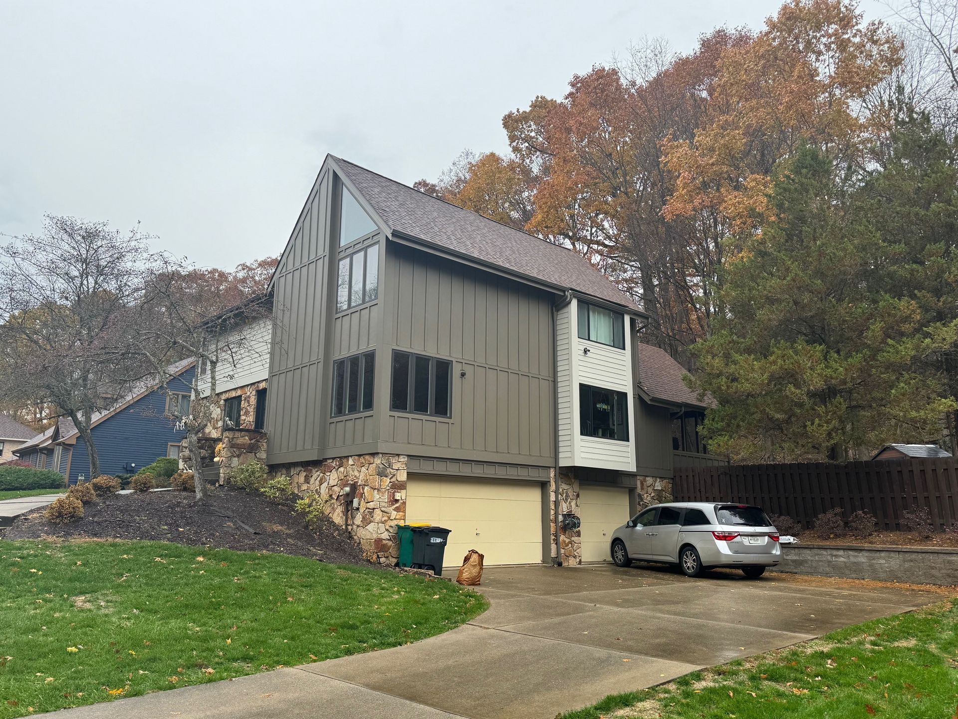 Two-story house with brown siding, stone accents, and a two-car garage. Car parked in driveway, dog on lawn. Autumn foliage.