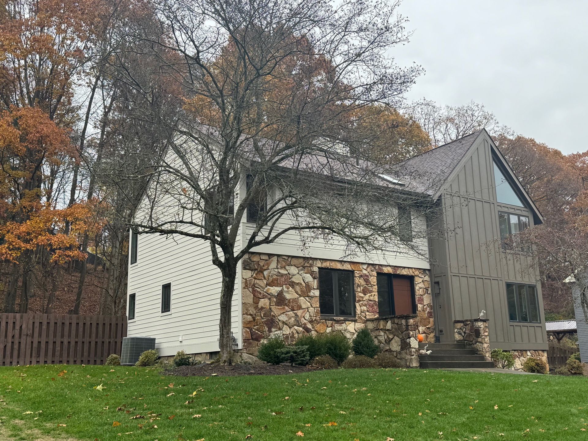 Two-story house with stone and tan siding, set in a yard with fall foliage and a brown fence.