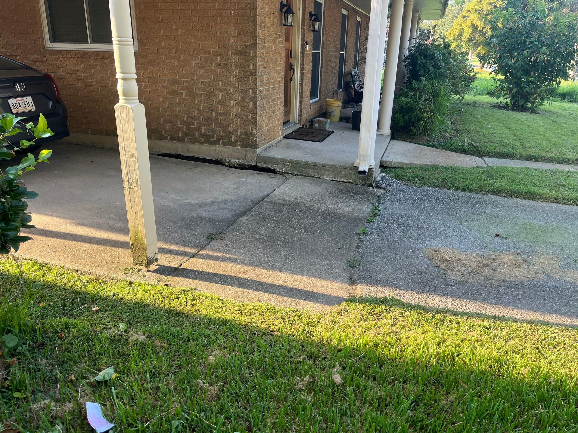 Cracked concrete path in front of a building with a porch, grass, and a parked car.