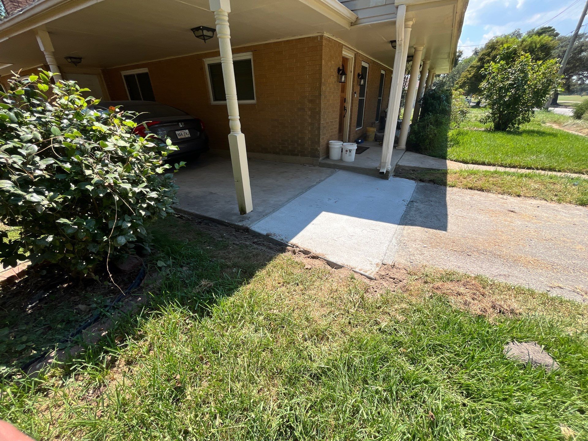 Carport with concrete path and grass in front of a brick building.